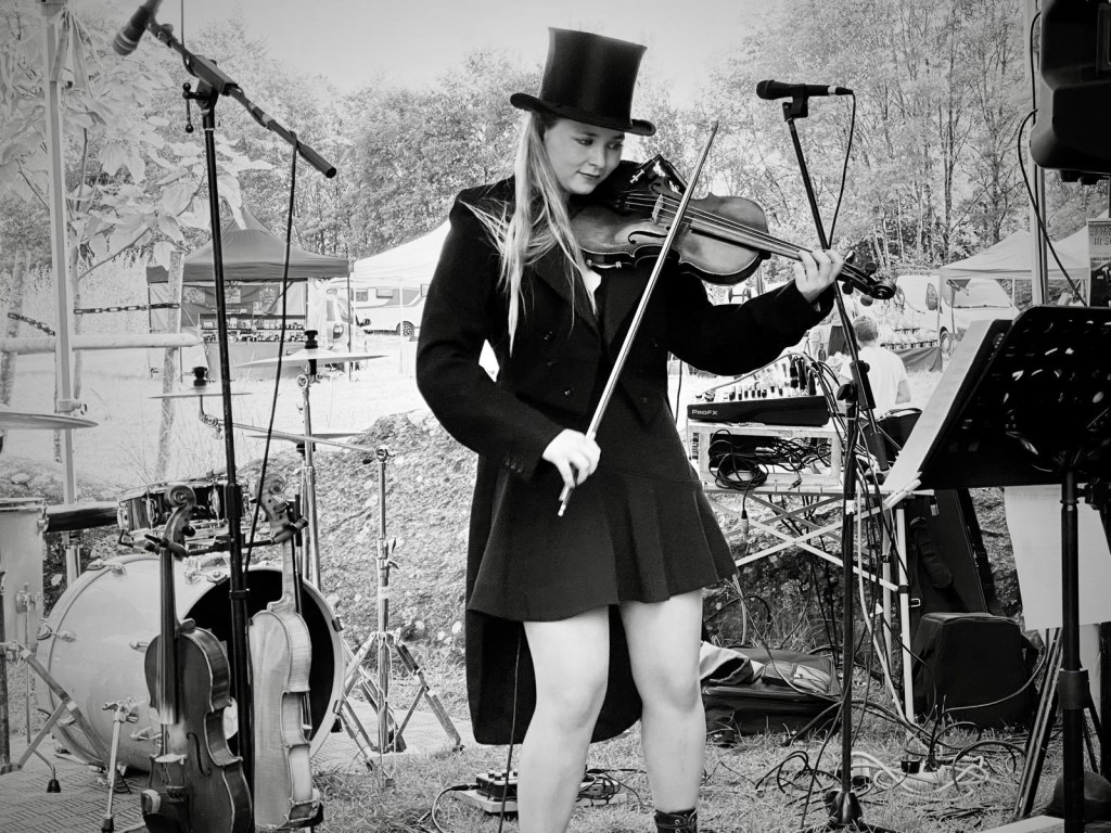 Violoniste en concert de plein air, vêtue d’un manteau noir et d’un haut-de-forme, jouant du violon sur scène avec énergie. Violoniste en Normandie lors d’un concert rock celtique, performance live avec violon et ambiance festive.