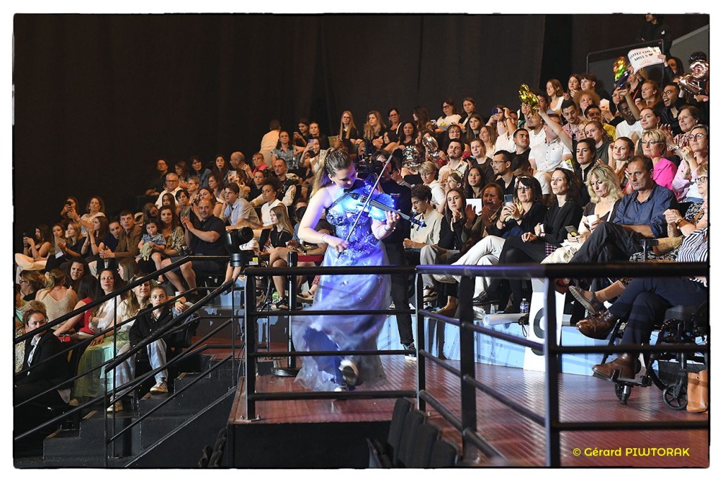 Violoniste jouant du violon électrique lumineux en robe bleue, se produisant au milieu du public d’une grande salle de spectacle. Show immersif de violon électrique pendant un événement live, violoniste jouant parmi le public pour une expérience unique.